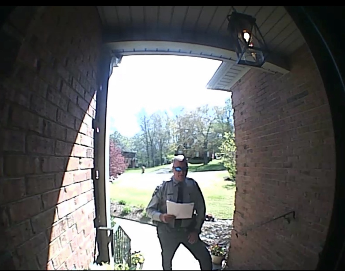 Doorbell-camera photograph of a uniformed officer at a residential front door, holding documents.